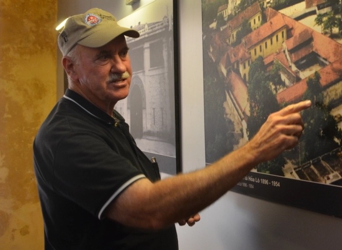 Older man in baseball cap pointing to a framed picture on the wall.