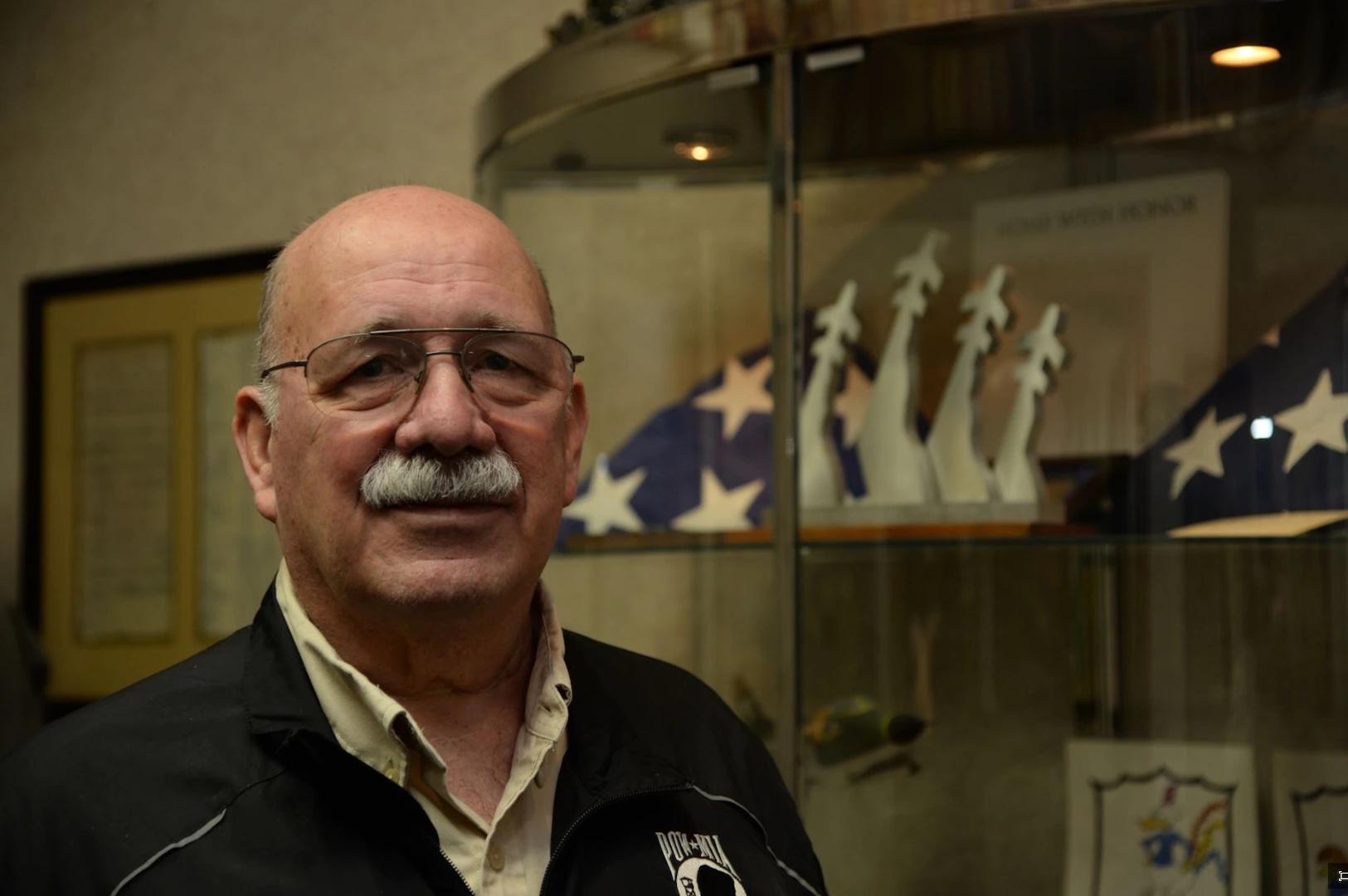 Man  in a jacket with a POW-MIA patch in front of a case of war memorabilia.