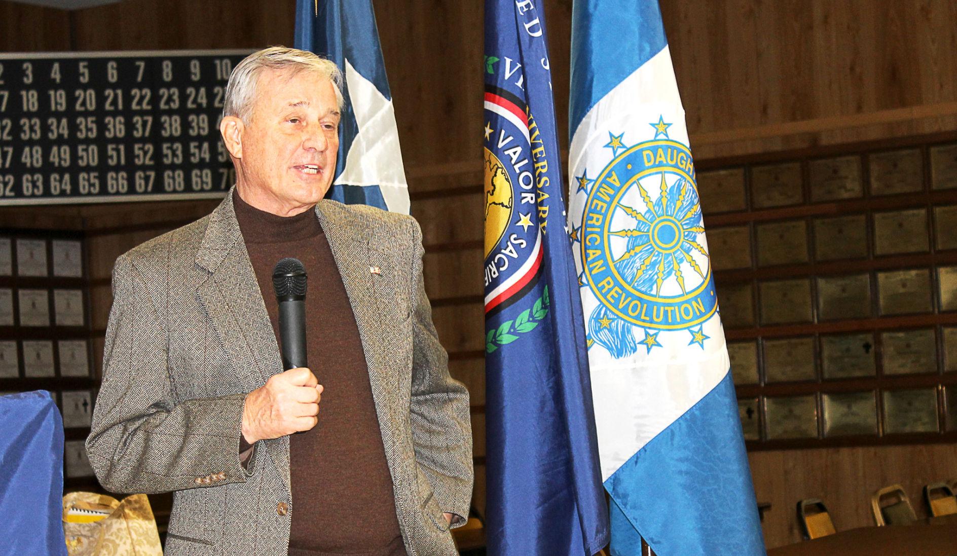 Man in a brown suit speaking into a microphone in front of flags and white and gold plaques.