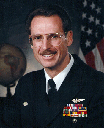 Man in a decorated U.S. Navy suit in front of an American flag and globe.