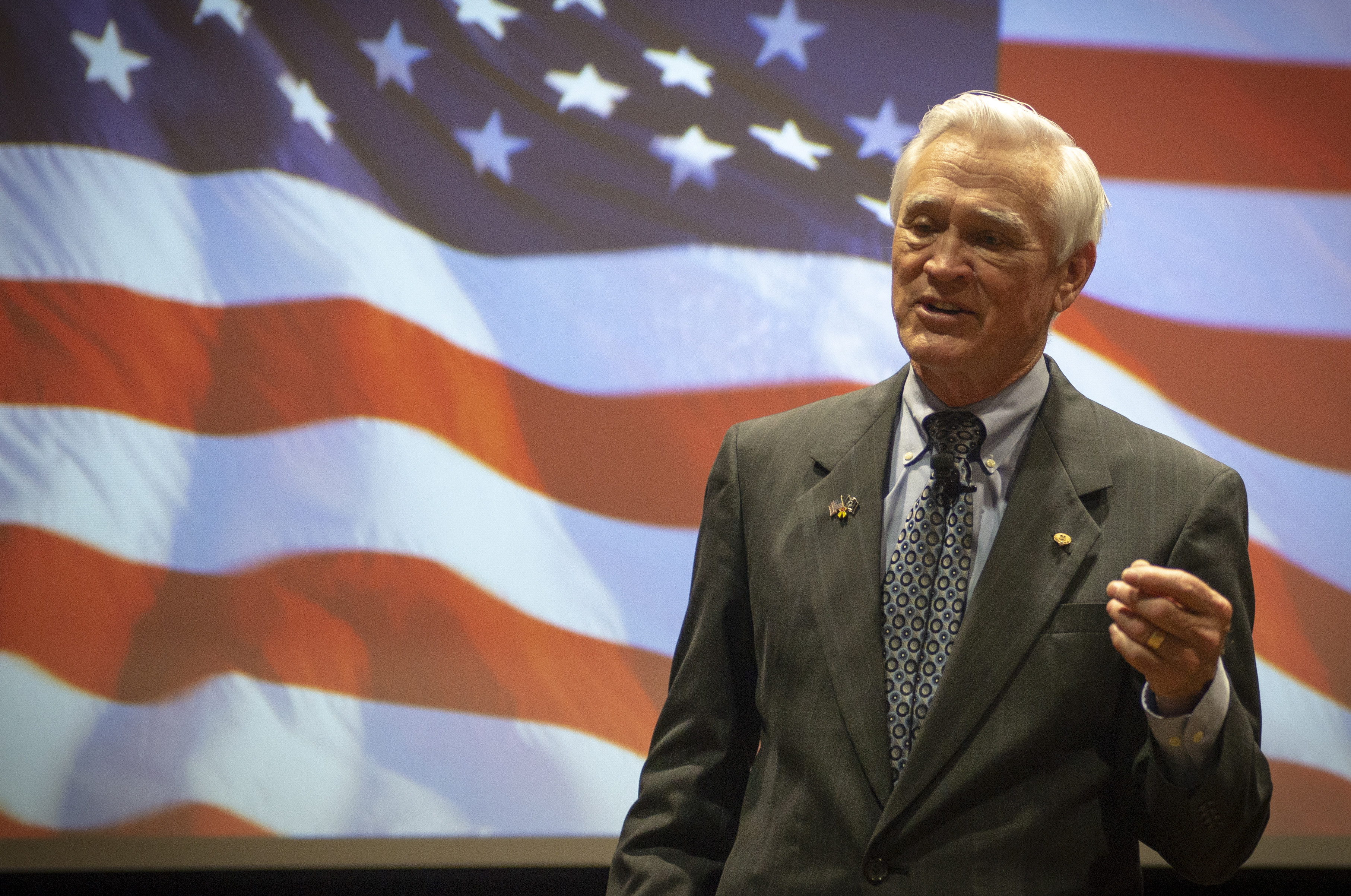 A man speaking in a suit and tie in front of a projection of an American flag.