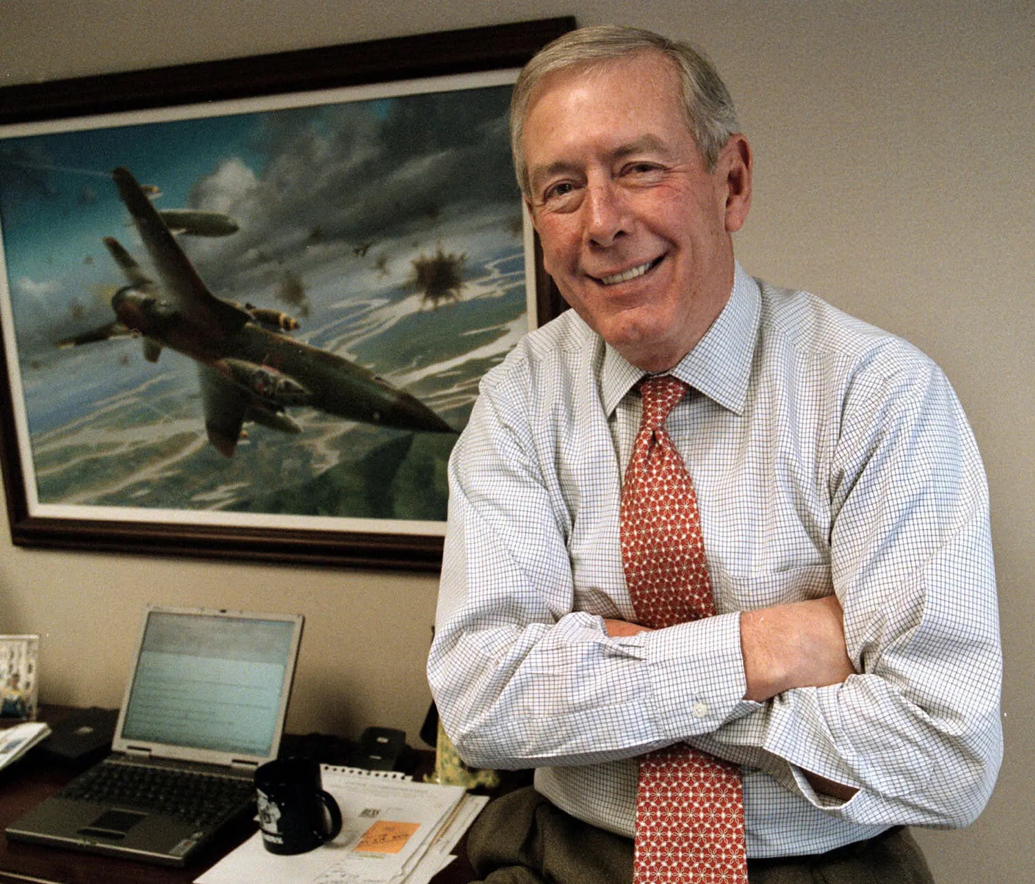 Older man with hands crossed sitting on a desk in front of a military plane painting.