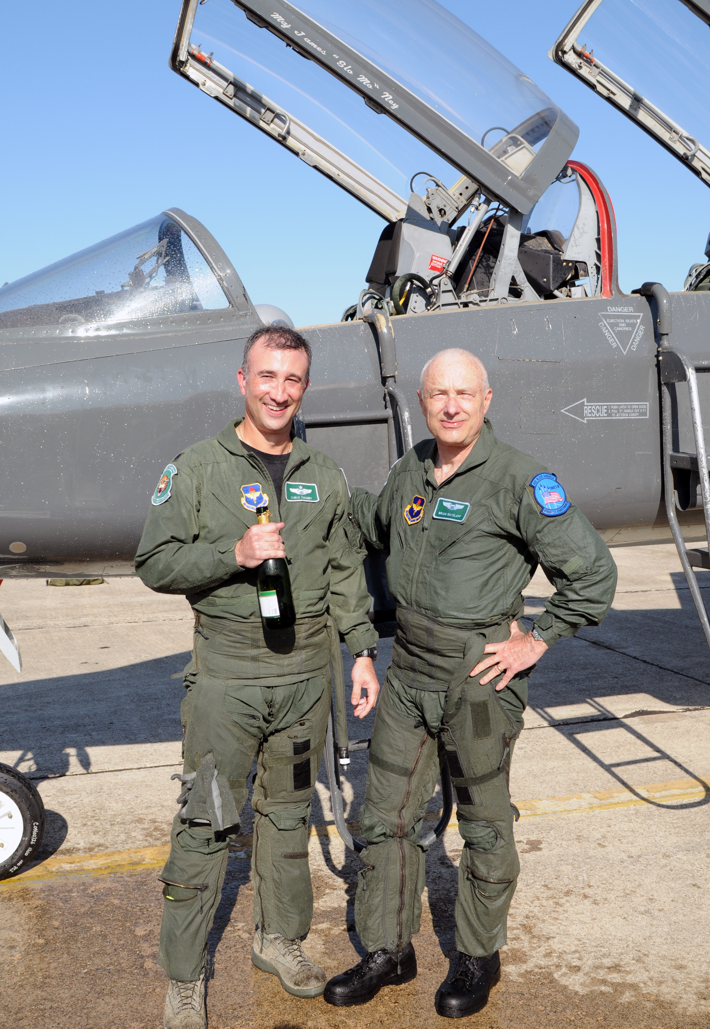 Two men in U.S. Air Force uniform smiling with a bottle of champagne in front of a military air craft.