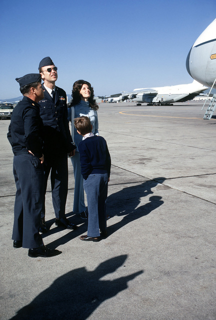 Two military officers standing with a woman and young boy on a runway with several aircrafts.