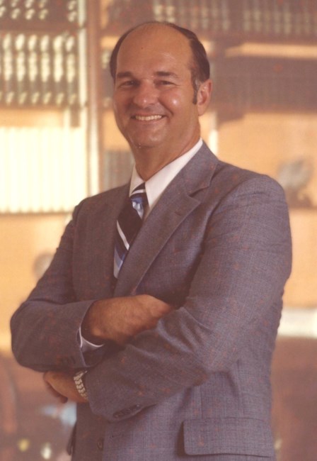 Man in a suit and tie smiling with his arms crossed in front of a bookcase.