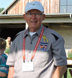 Man in a grey collared shirt, a pinned yellow ribbon, red lanyard and white hat.