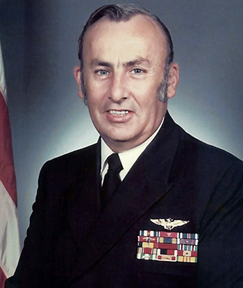 Man in a decorated U.S. Navy suit next to an American flag.