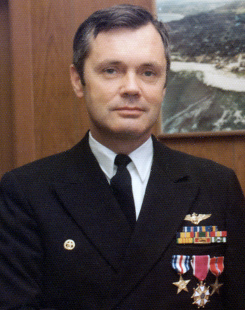 Man in a decorated U.S. Navy suit standing in front of a wooden wall and photograph.