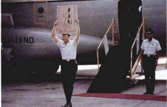 Man walking off aircraft smiling and holding a sign with a fellow officer behind him.