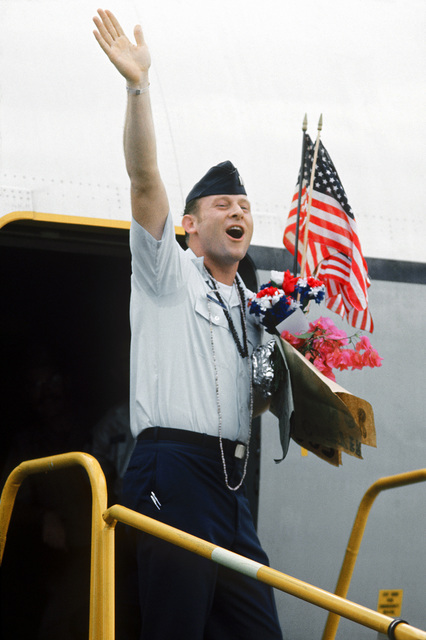 Man in a U.S. Air Force uniform waving while leaving an aircraft with a bouquet of flowers and American flags in his hands.