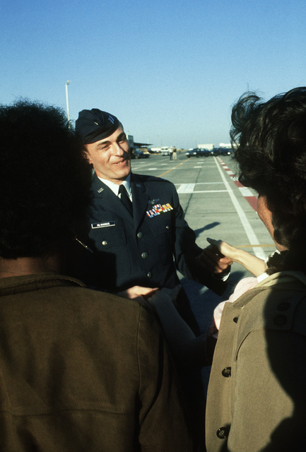 Man in military uniform and cap talking to a crowd on an aircraft runway.