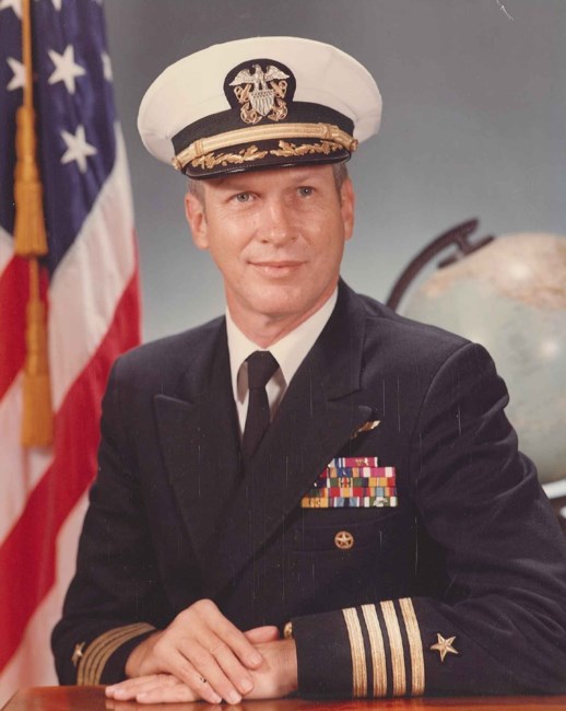 Man in a decorated U.S. Navy suit and cap in front of an American flag and world globe.