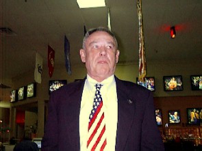 Man in a suit with an American flag tie in front of a wall of photographs and flags.