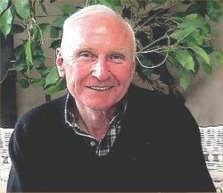 Older man smiling in a collared shirt and sweater in front of a plant.