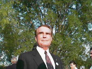 Man in a suit a tie in front of foliage.