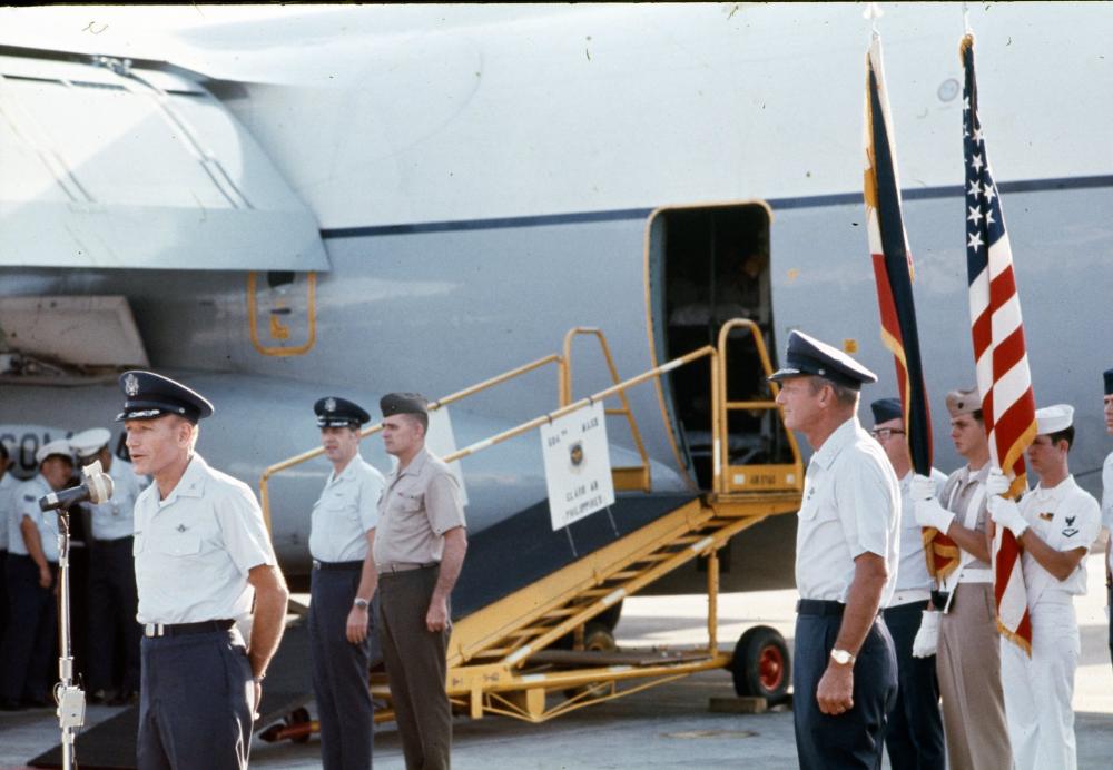 Multiple officers standing in front of an aircraft. One officer speaking into a microphone.