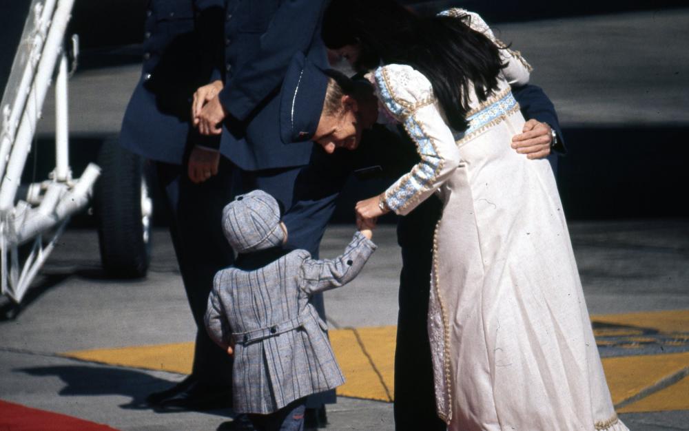 Officer on tarmac leaning down to talk to young child while embracing a woman.