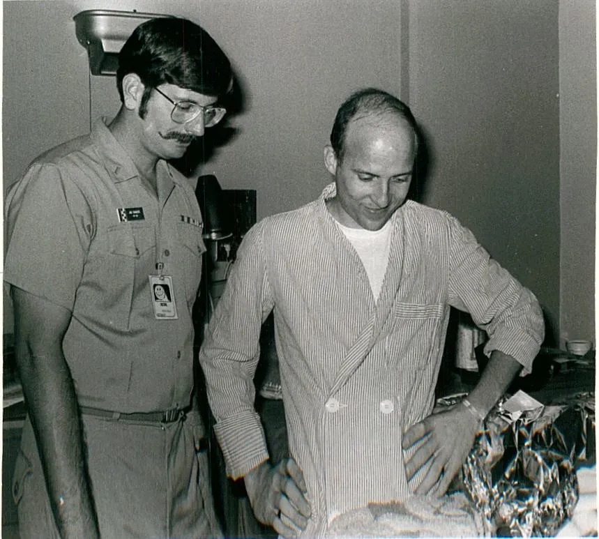 Two men, one a military officer uniform and the other in jacket ,standing, gazing at something on a table.