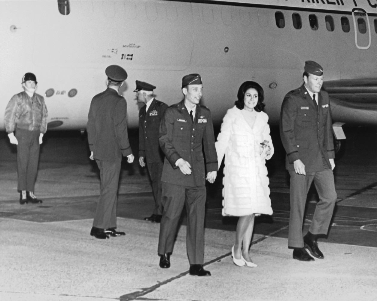 Men in military uniform on a tarmac in front of an aircraft. One man walking holding hands with a woman in a fur coat.