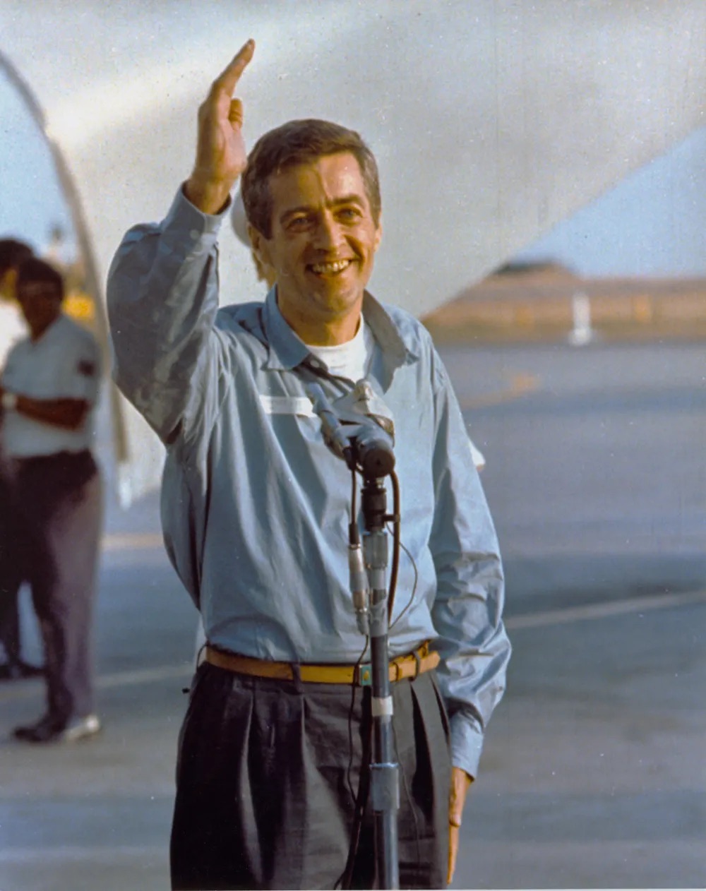 Man standing at a microphone on a tarmac with his hand raised.