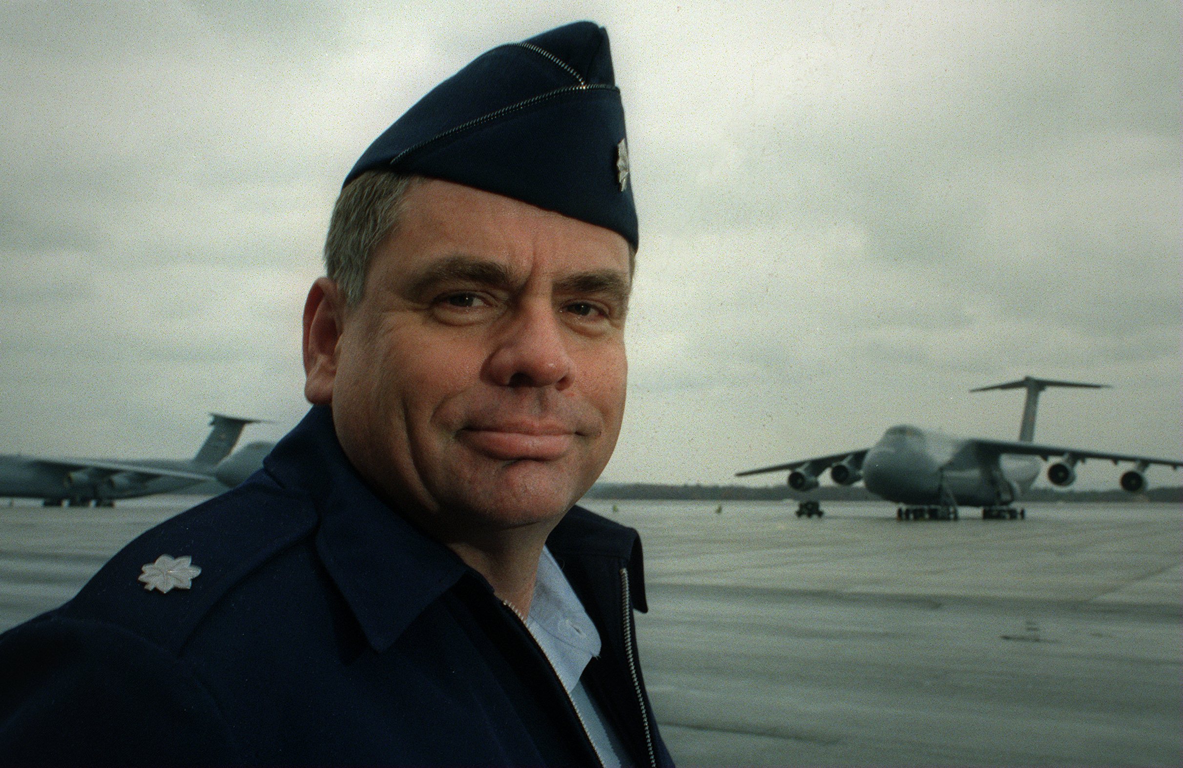 Man in a military suit and cap on a tarmac with aircrafts in the background.