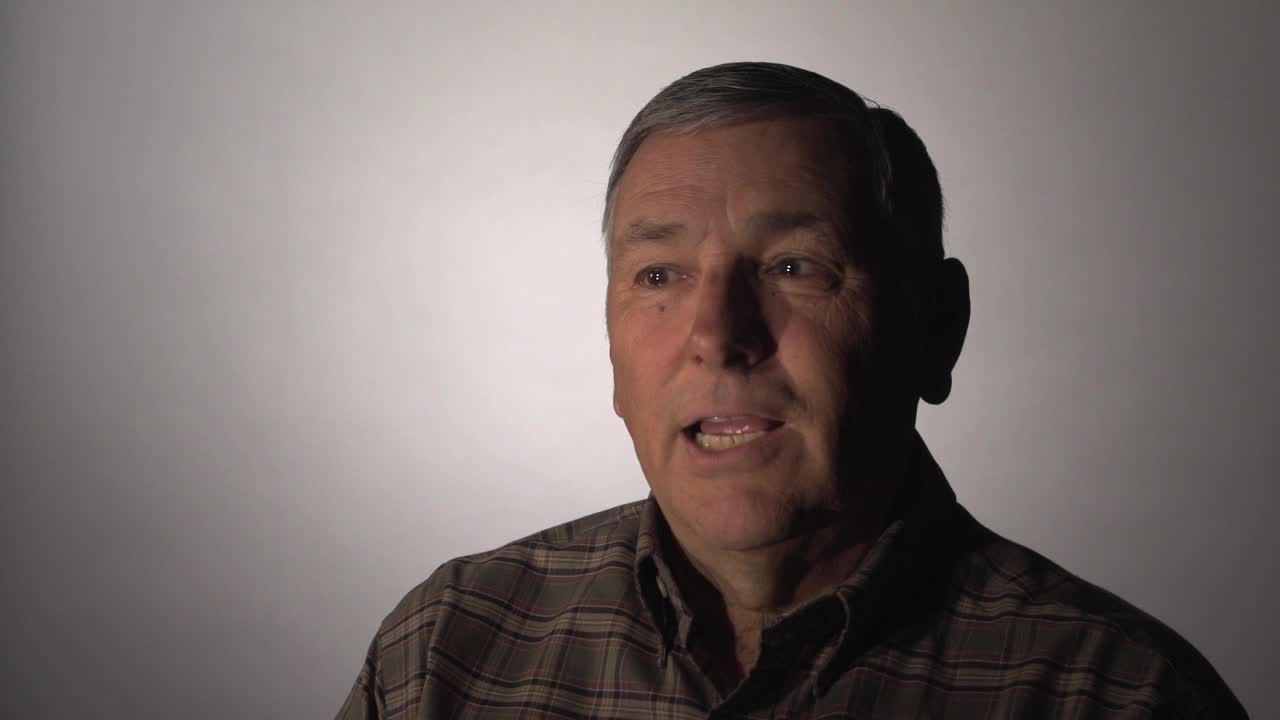 Man in a plaid shirt speaking in front of a white background.