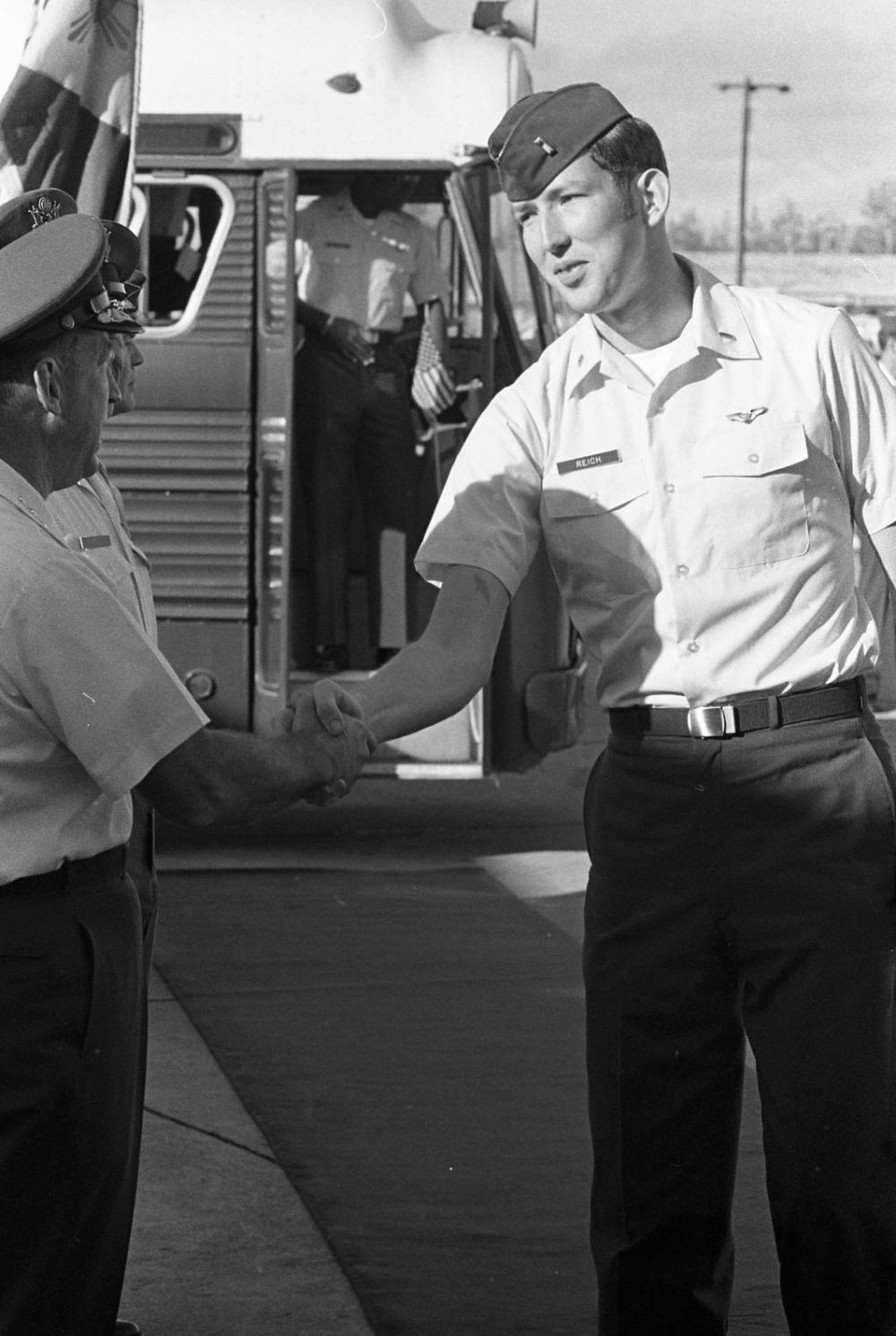 Military officers shaking hands on a carpet in front of a bus on a tarmac.