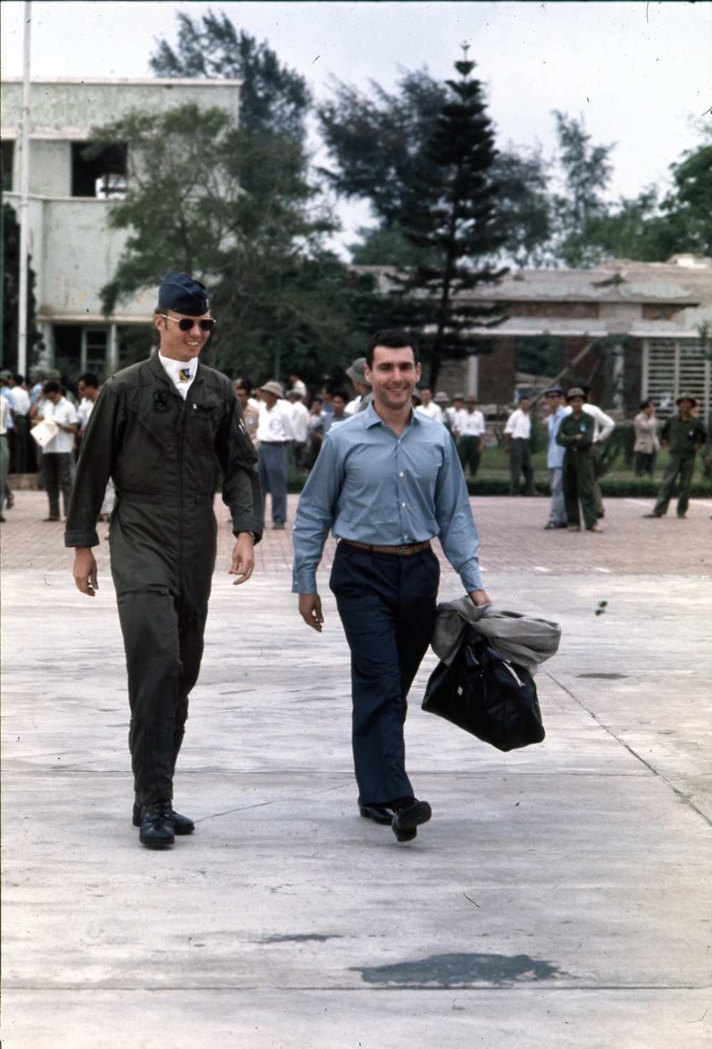 Man in a blue button down a pants carrying bags on a tarmac next to a military officer in front of a crowd.