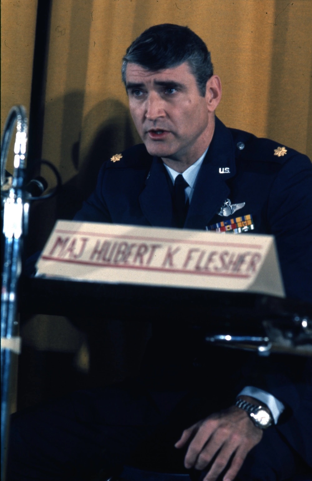 Man sitting in front of a yellow curtain speaking with a name tag with "MAJ Hubert K. Flesher" in the foreground.