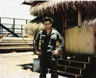 Man standing in front of a hut in a military uniform.