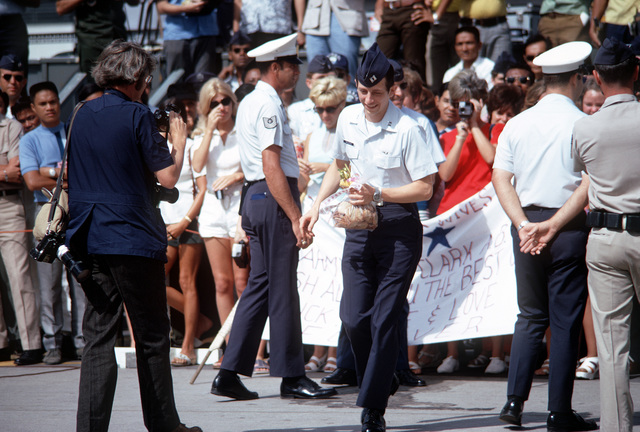 A man in military uniform walking with a bag in his hand in front of fellow officers and a large crowd with signs and cameras.