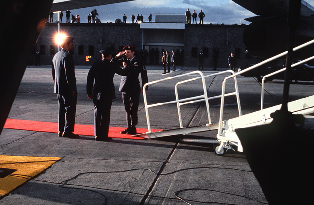 Man on a red carpet laid in front of an aircraft ramp saluting fellow officers.